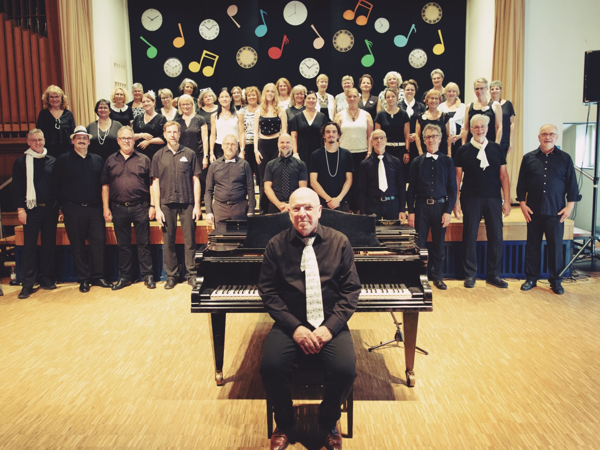 Lothar Sigloch sitzt vor einem Klavier. Hinter ihm steht der Chor MET der Liederlust Mettingen für ein Gruppenfoto auf der Bühne.